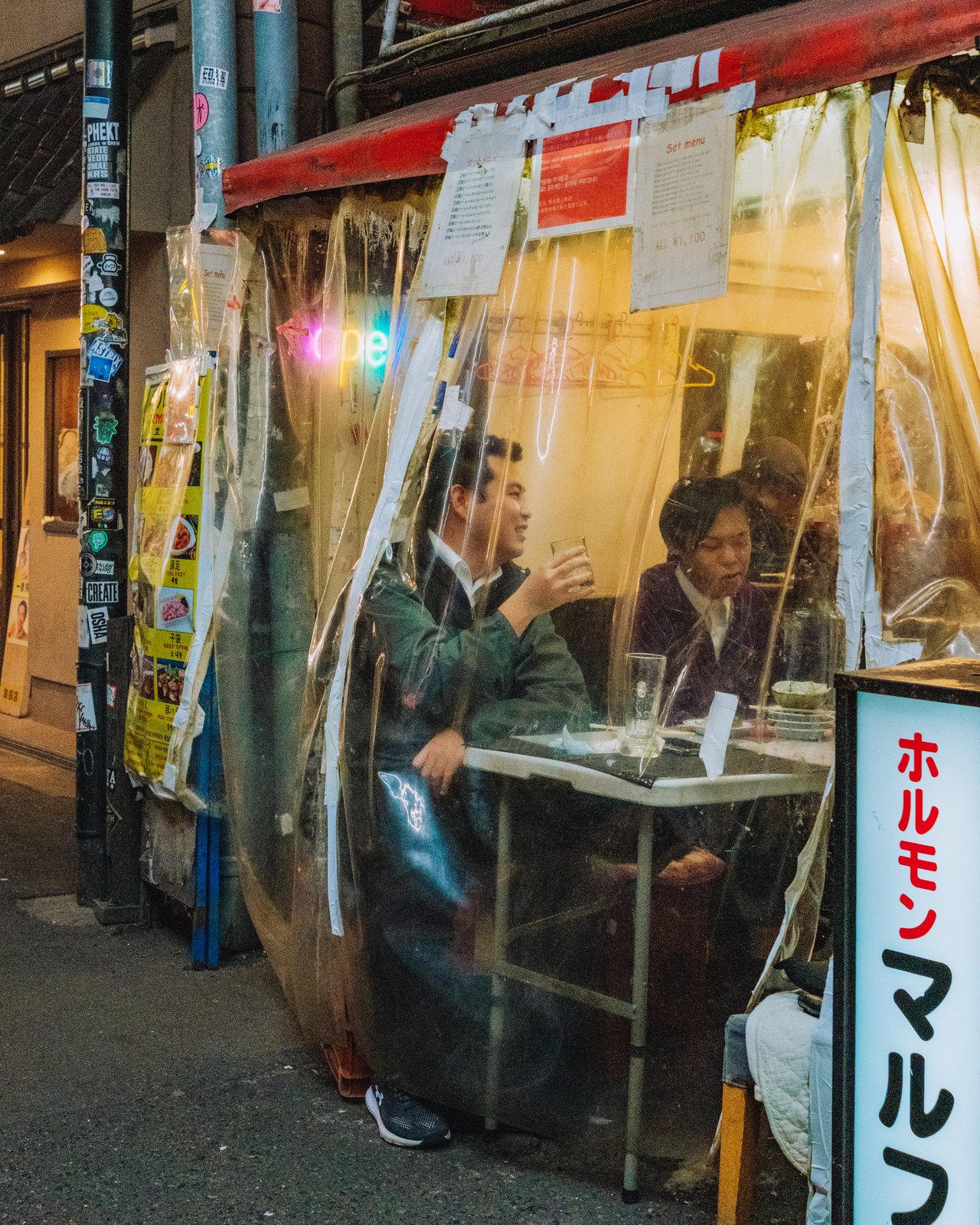 Two men drinking at a tiny plastic-curtained street stall at night, Osaka