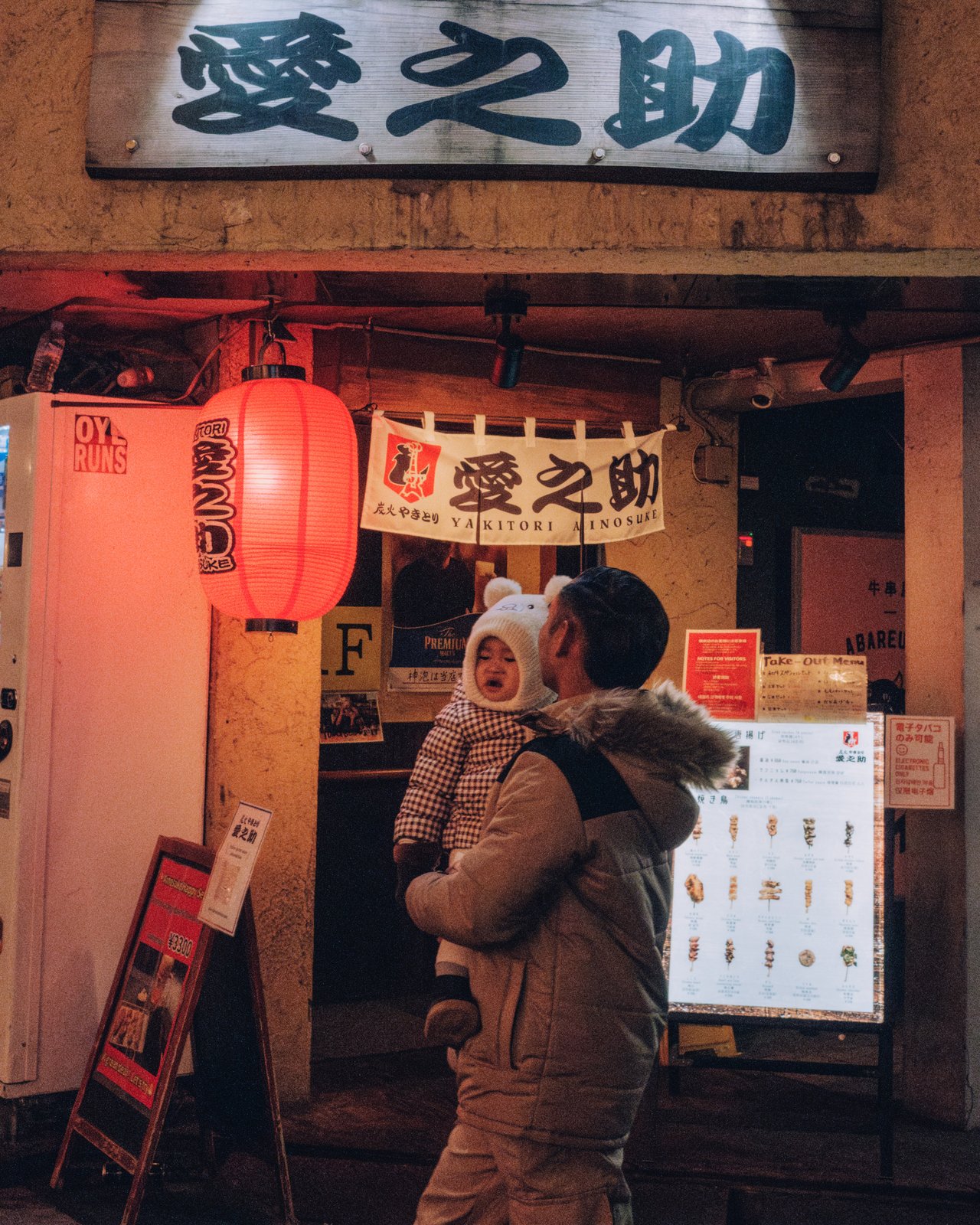 A man holding a small child outside a lantern-lit yakitori restaurant at night, Osaka