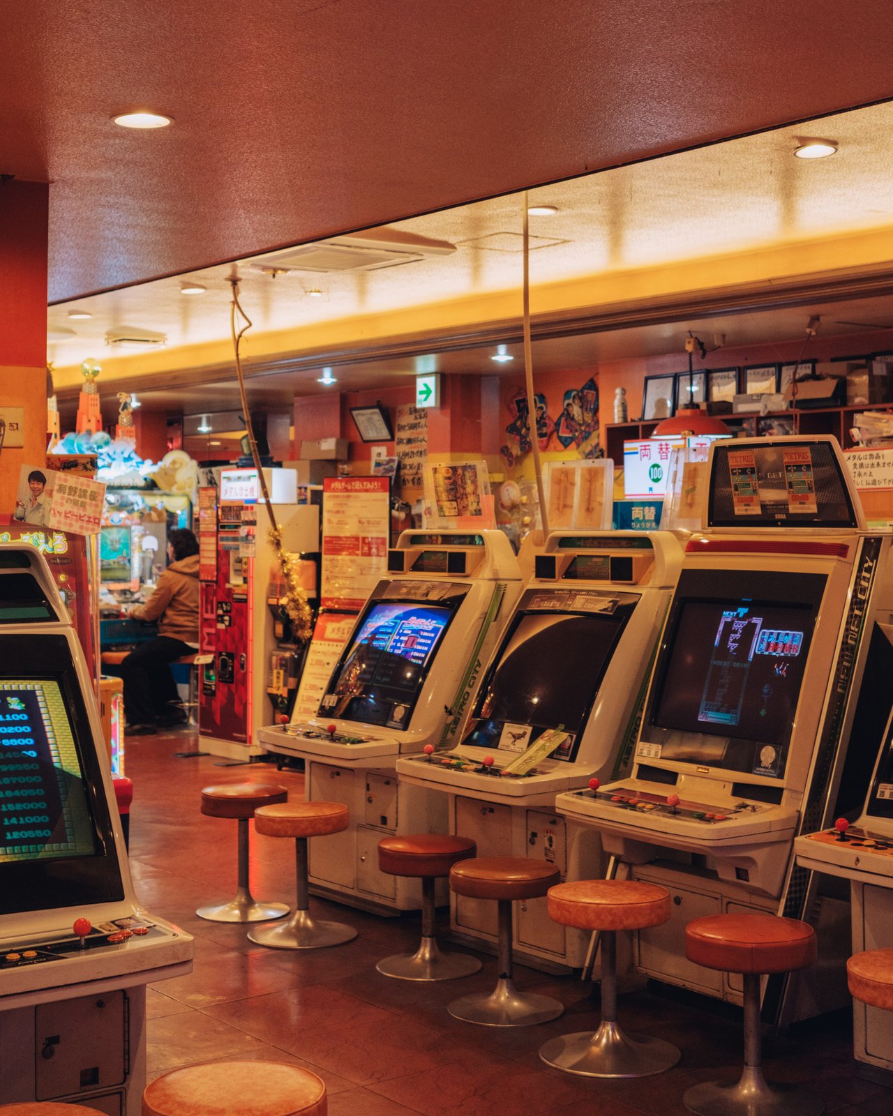 A retro arcade with a row of vintage sit-down cabinets and orange stools, Osaka