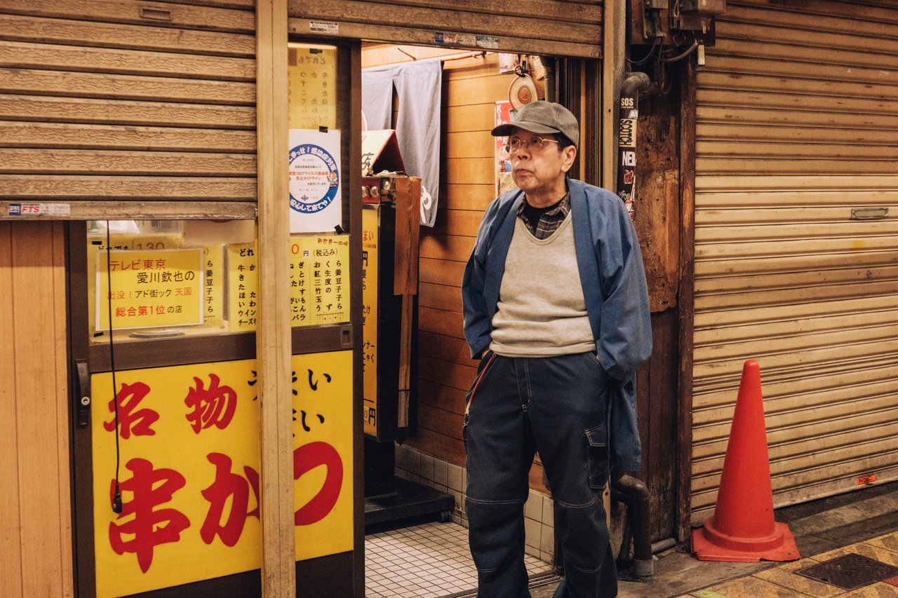 An older man standing by a shuttered shop and a kushikatsu banner at night, Osaka