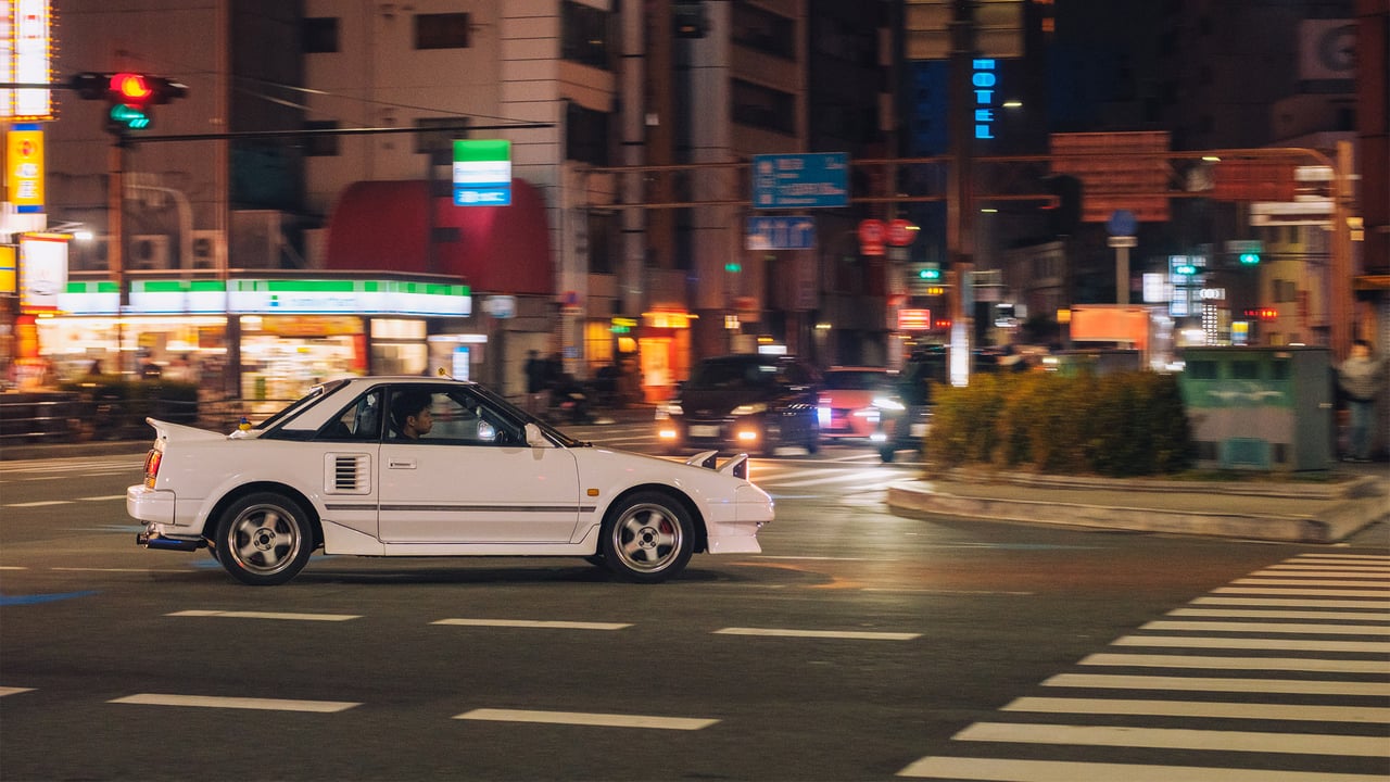 A white classic sports car crossing a neon-lit intersection at night, Osaka, Japan