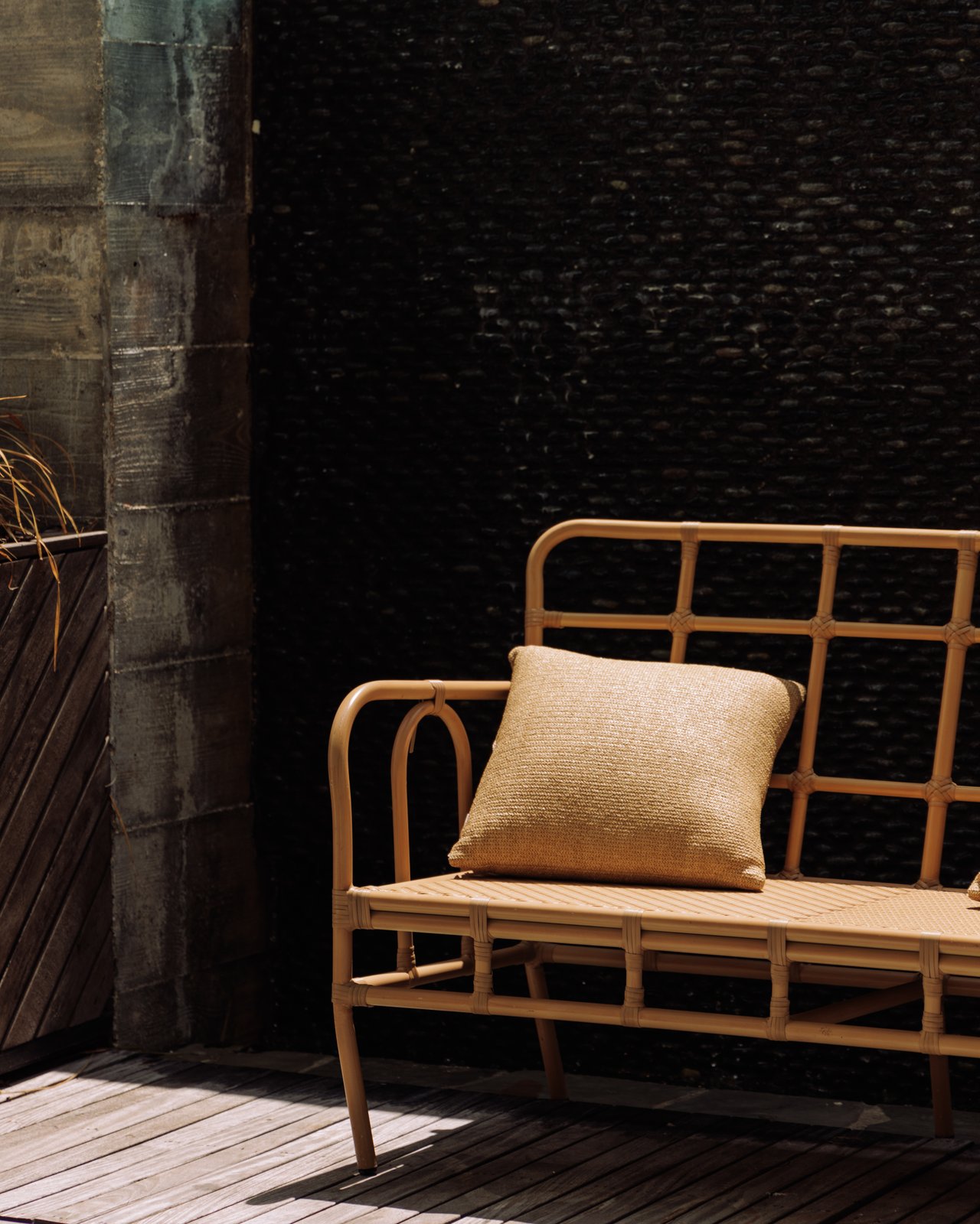 A cane settee with a cushion on a wood deck against a dark stone wall, The Balfour Hotel