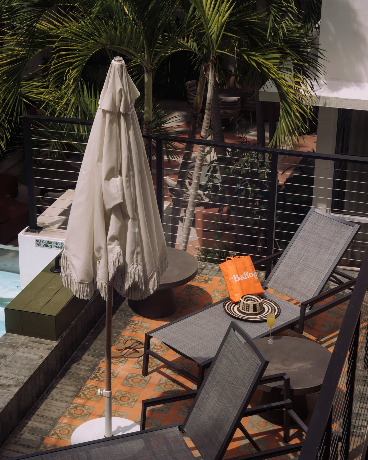 A poolside deck with grey chaises, a fringed umbrella and an orange Balfour tote and sun hat, The Balfour Hotel