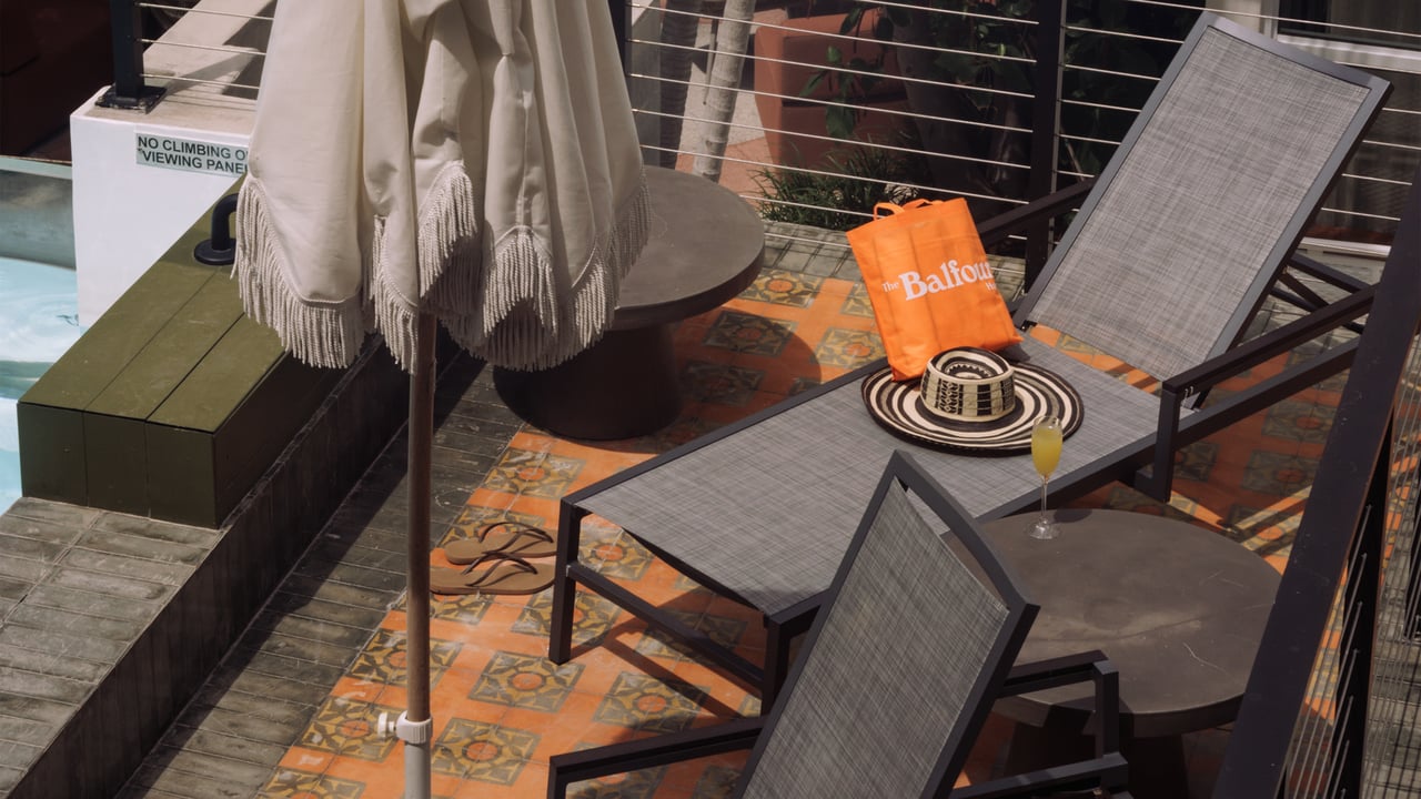 A poolside terrace with grey chaise lounges, a fringed umbrella and an orange Balfour tote and sun hat, The Balfour Hotel, Miami Beach