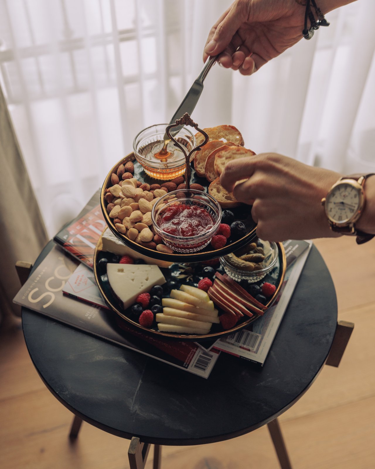 A hand spreading jam on toast over a tiered breakfast board of nuts, cheese and fruit on a marble bistro table