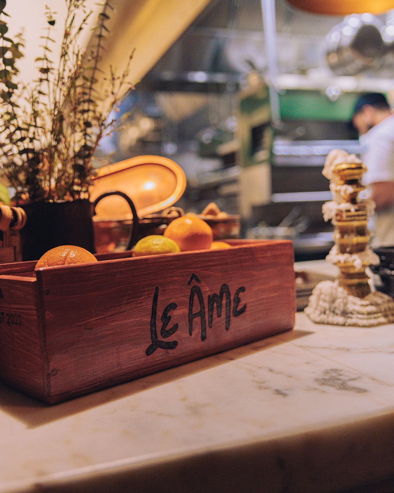 A wood crate of oranges marked 'Le Âme' on a marble counter beside the open kitchen