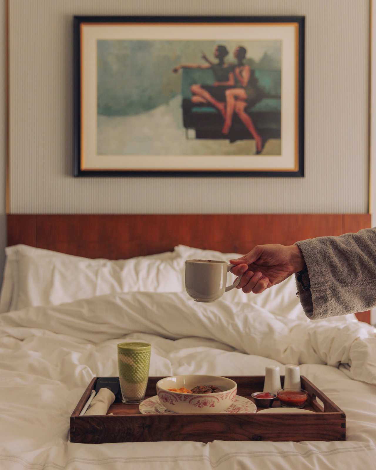 Breakfast in bed — a hand lifts a coffee cup from a wood tray on white linens