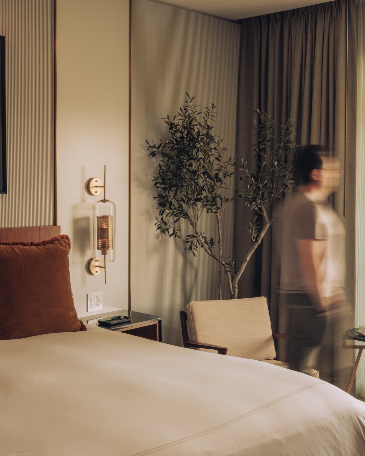 A guest room at dusk, a figure passing in motion blur beside the bed and a potted tree