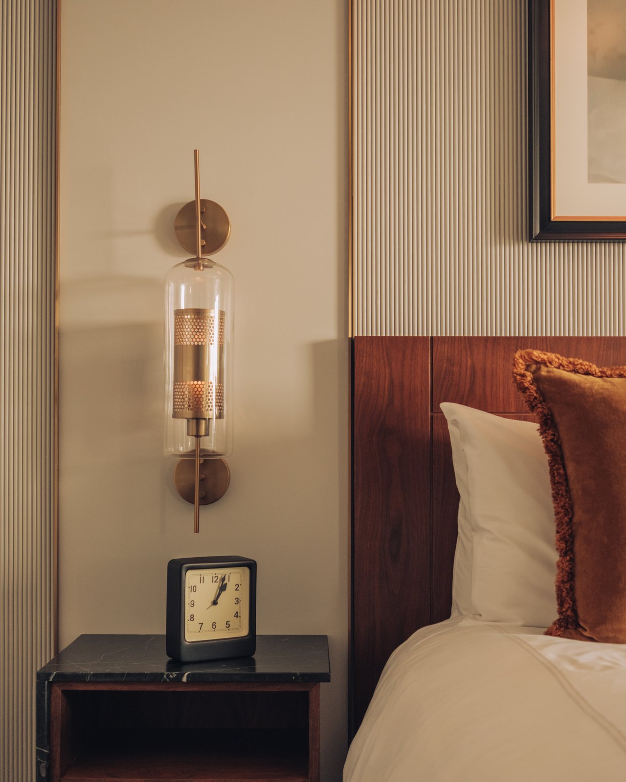 A bedside detail — a glass-and-brass sconce and a small square clock on the nightstand