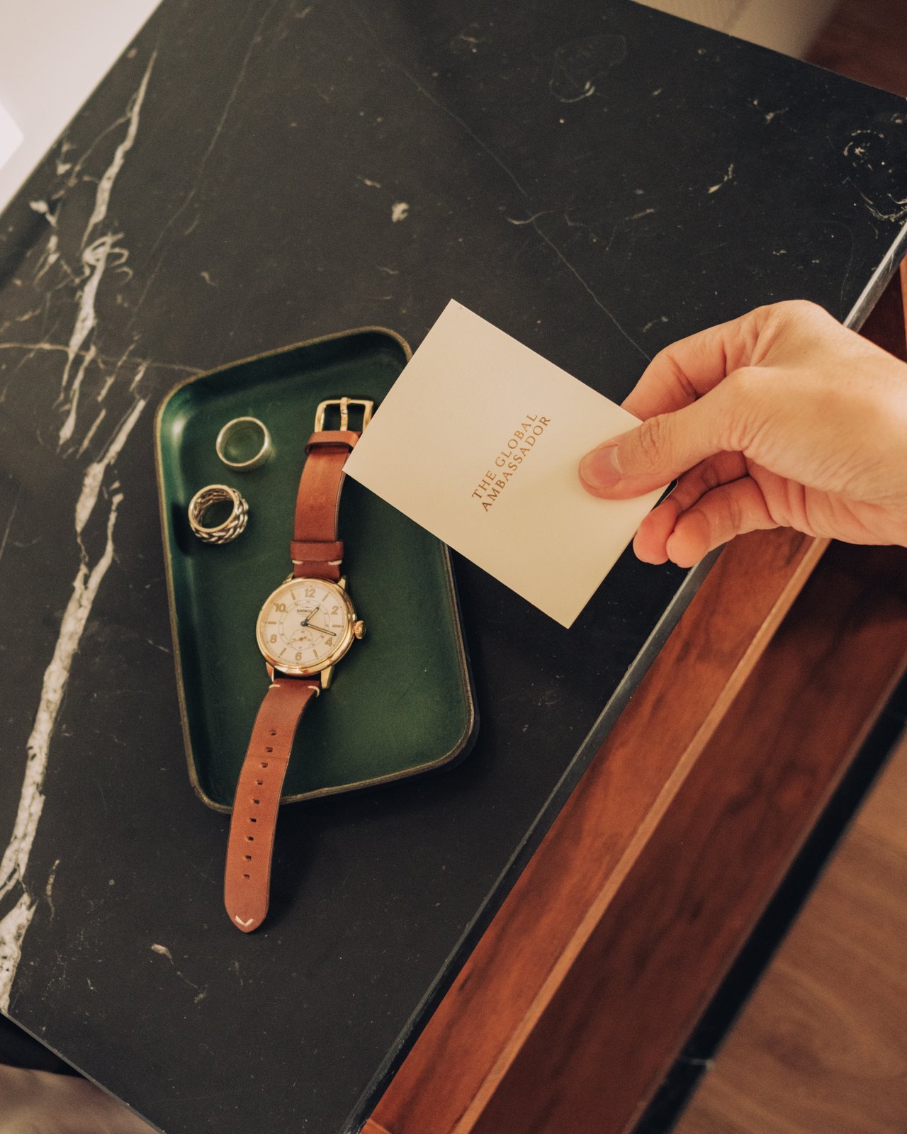 A hand setting a card on a green leather valet tray holding a watch and rings at The Global Ambassador
