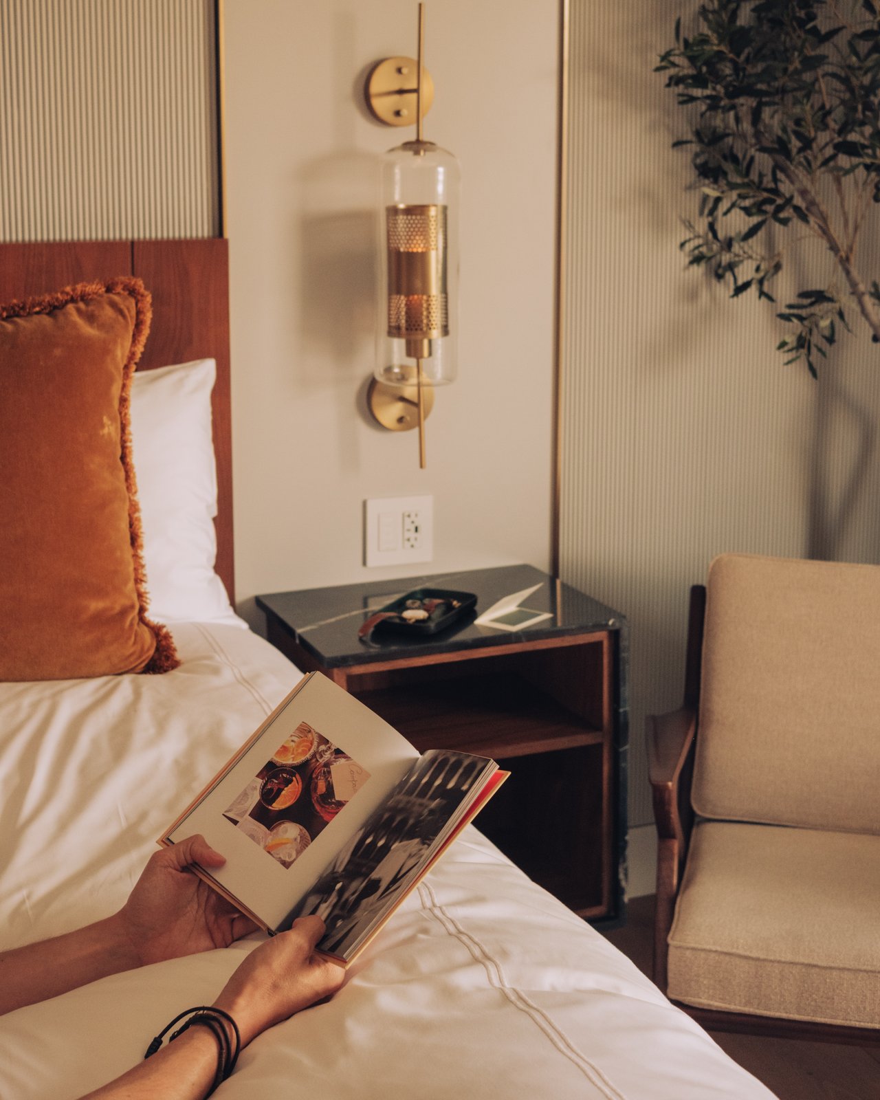 A guest reading in bed, a wall sconce lit and an armchair alongside