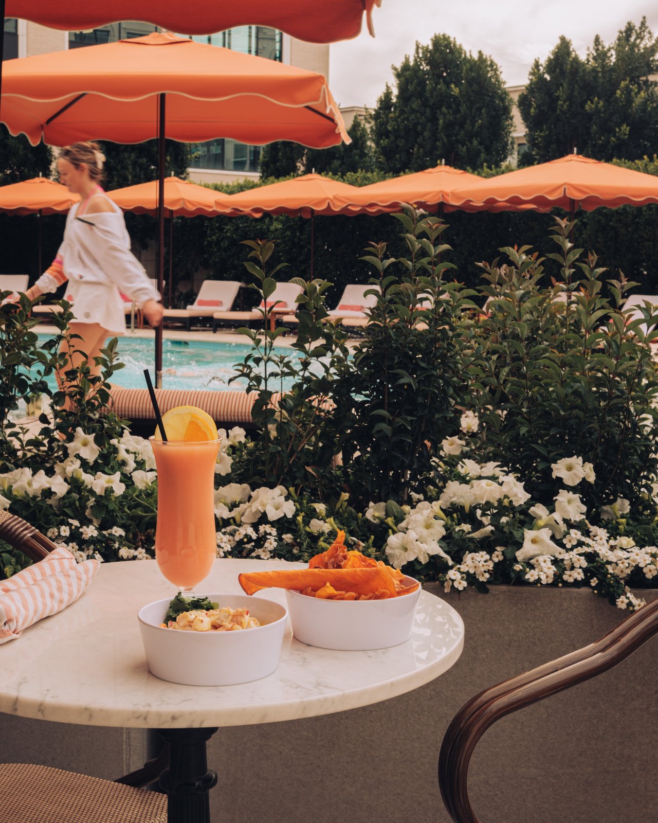 A poolside marble table with a bright orange drink and a snack, white flowers and the pool behind
