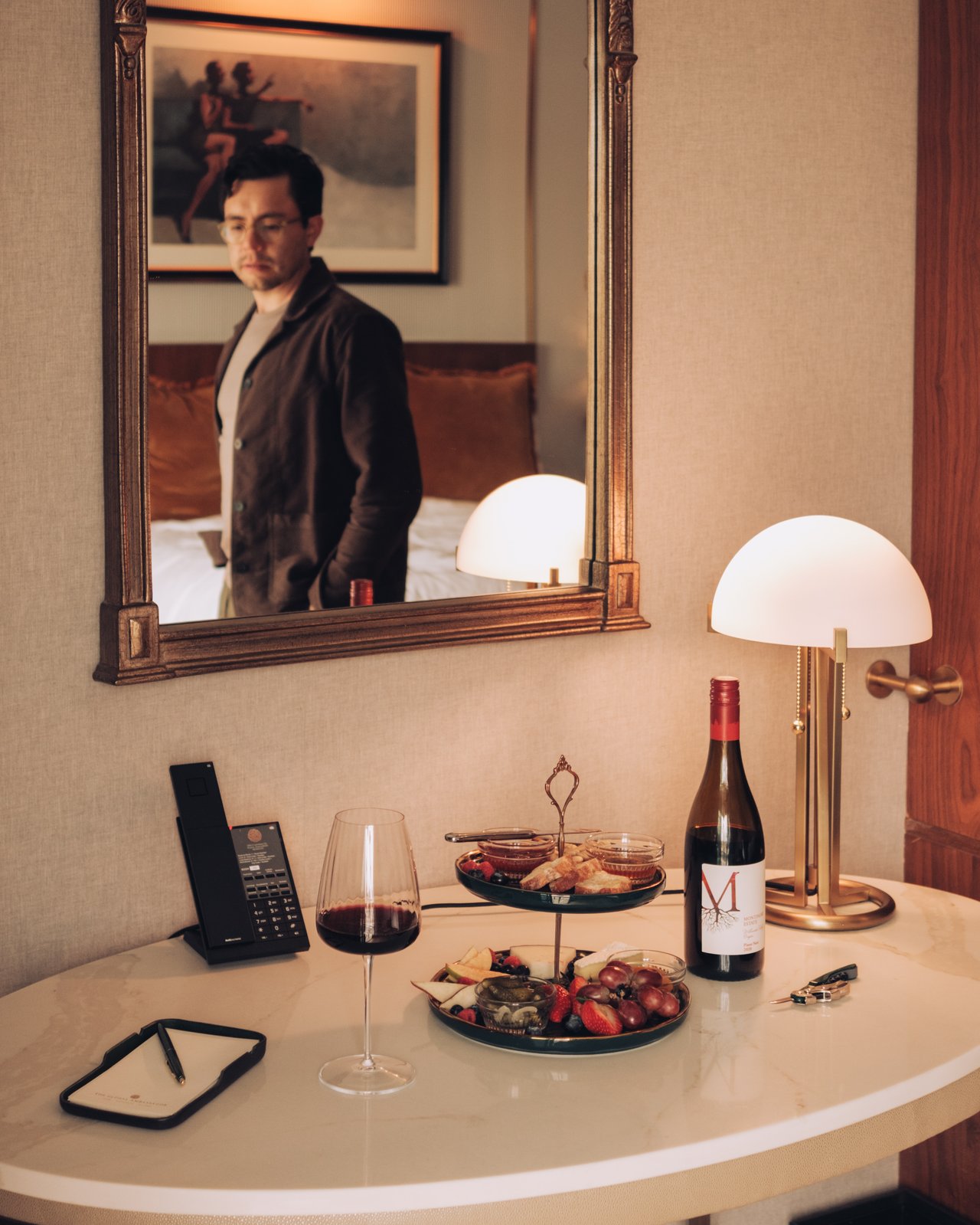 A guest-room console with wine, a tiered snack tray and a mushroom lamp, a man reflected in the mirror
