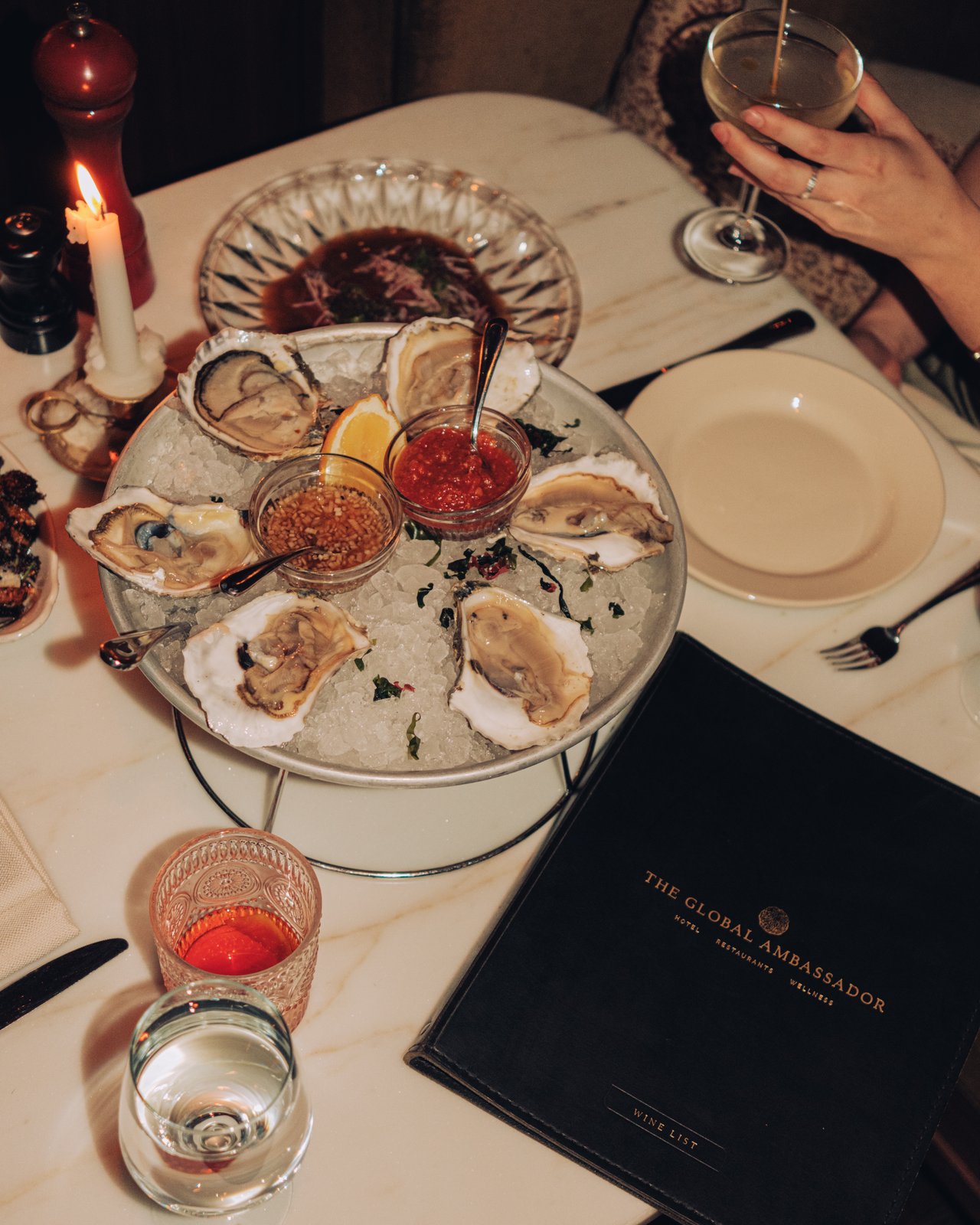 A raised seafood tower of oysters on ice with sauces, a candle and a martini on marble