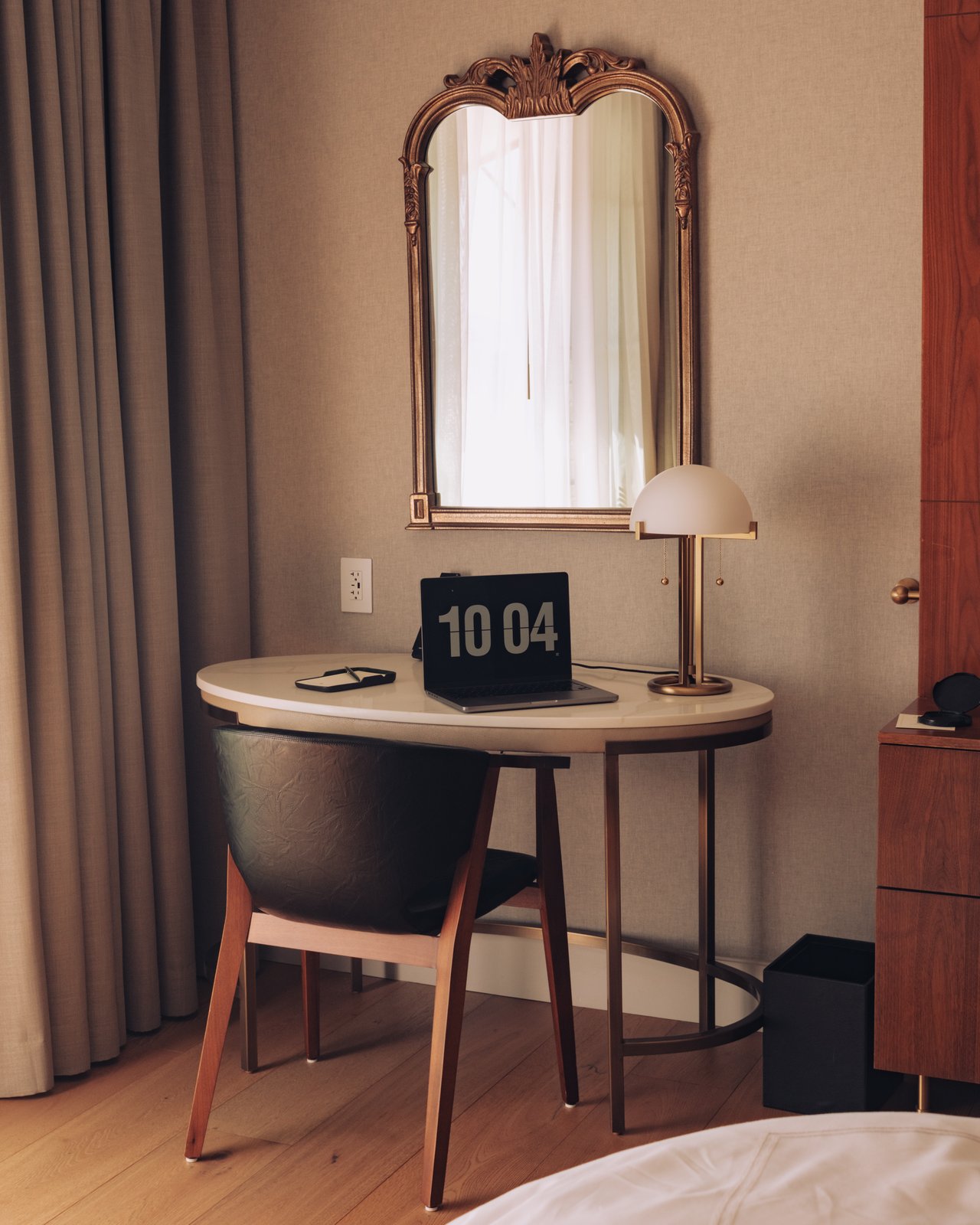 A round writing desk with a flip clock reading 10:04 beneath an arched gilt mirror