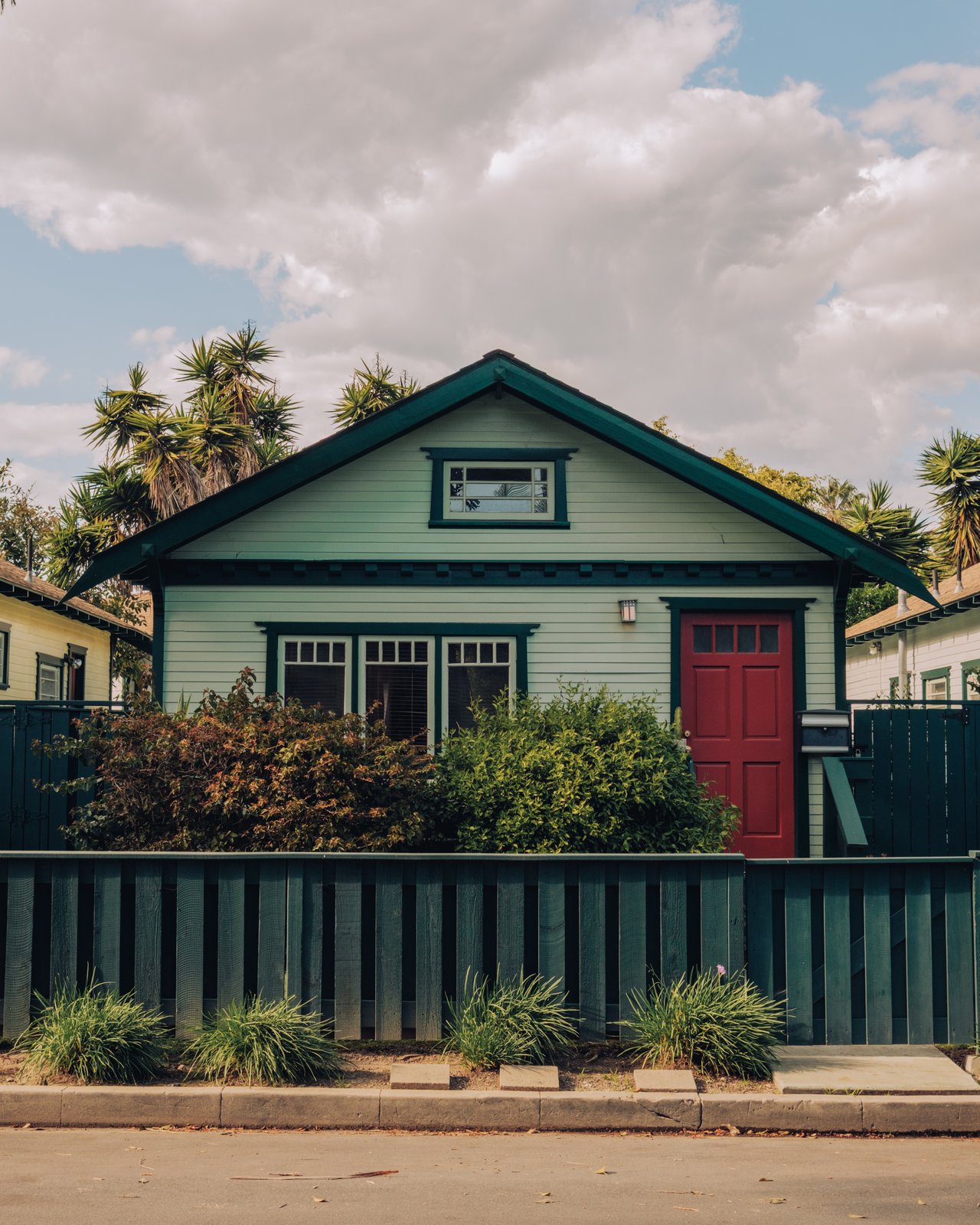 A teal Craftsman bungalow with a red door behind a green fence, Venice