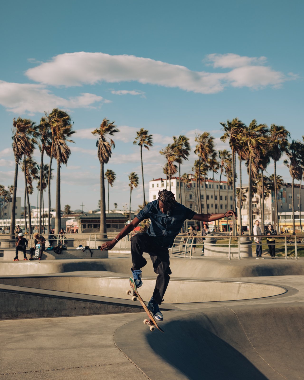 A skateboarder mid-trick at the Venice skate park, palms and boardwalk behind