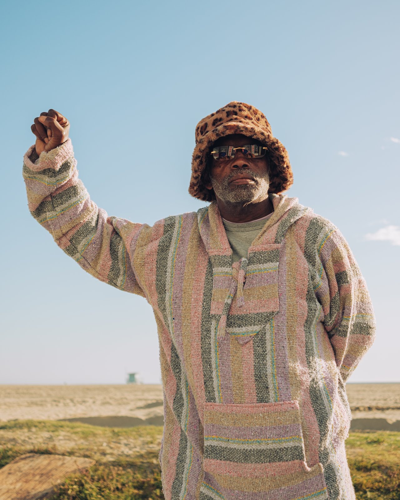A man in a Baja hoodie and leopard hat raising his hand, a portrait at Venice Beach