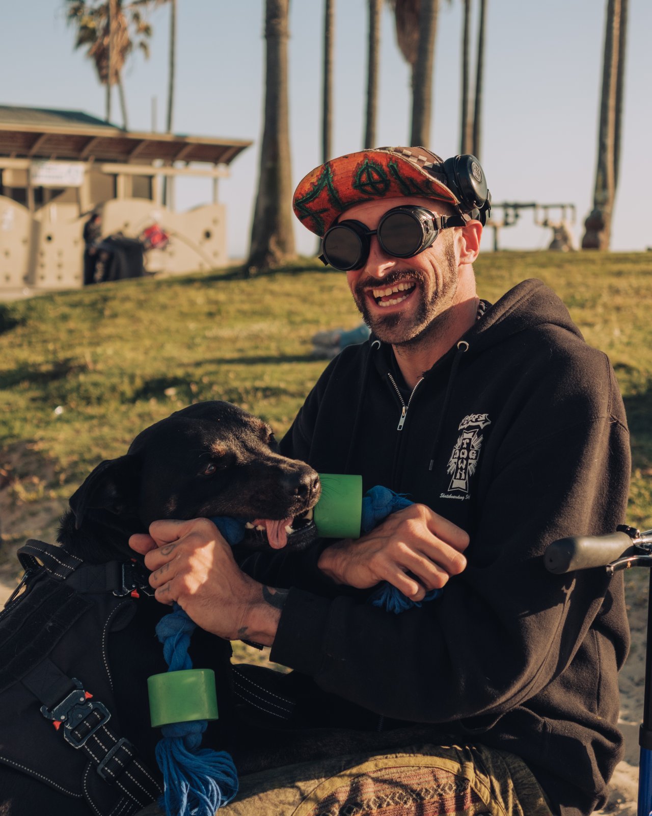 A grinning man in goggles holding his black dog on the grass, Venice