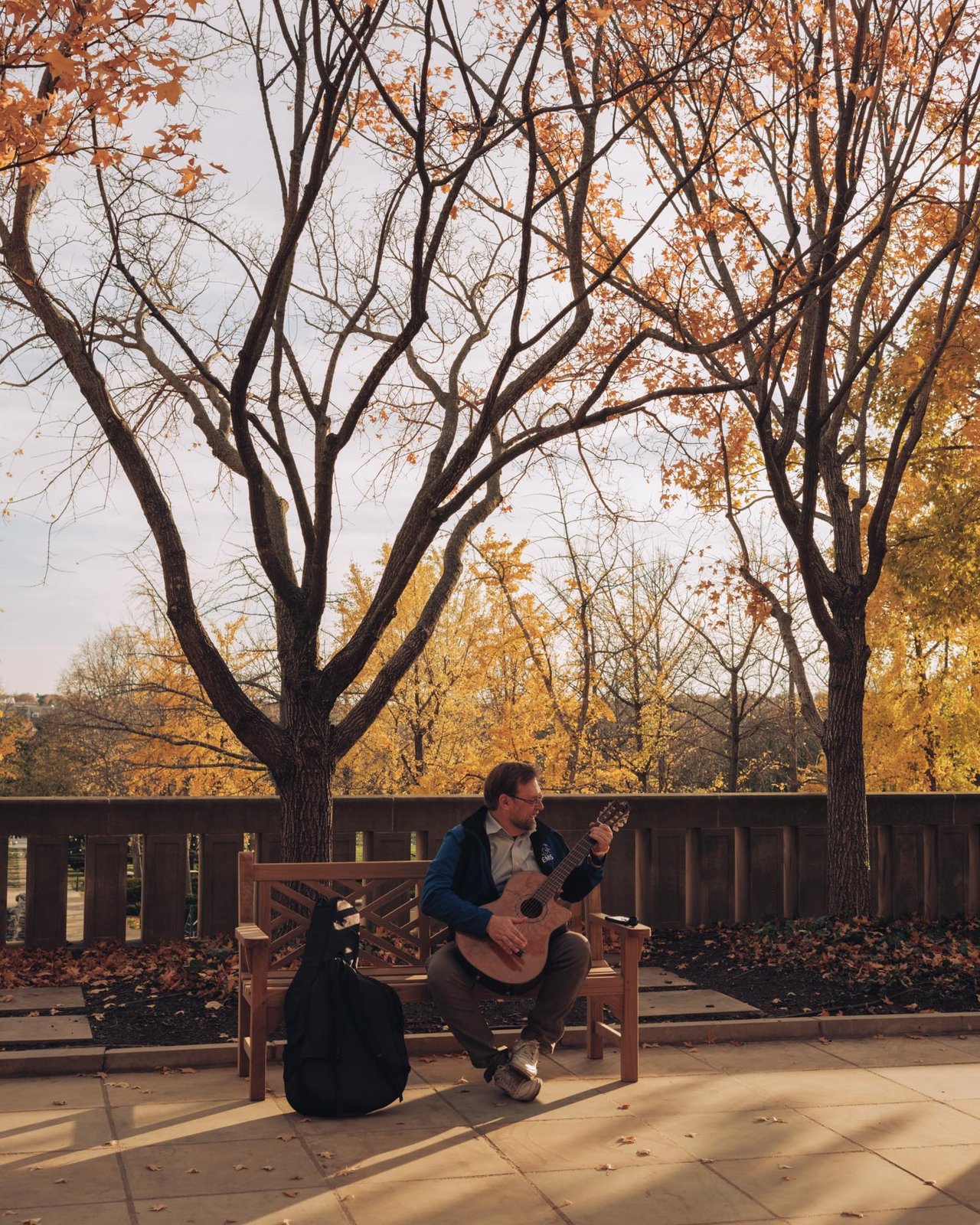 A man playing guitar on a bench under bare autumn trees, Kansas City