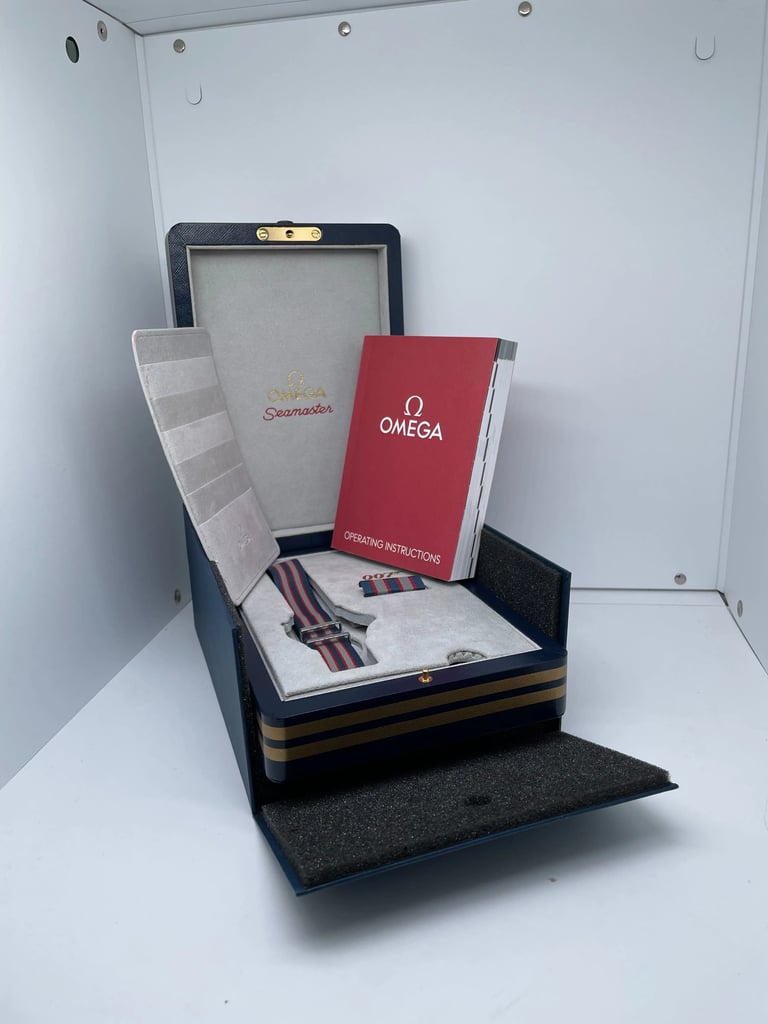 An image of a pre-owned men's OMEGA wristwatch displayed inside an open box. The watch features a striped fabric strap, and the box is situated at a slight angle in the foreground. A red booklet labeled "OMEGA" and "Operating Instructions" is visible, leaning against the inner lid of the box. The watch is positioned at a medium distance, providing a clear view of the packaging and accessories.