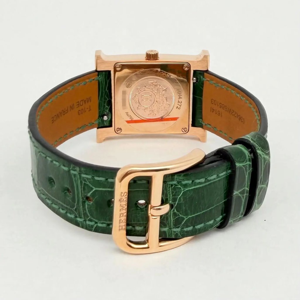 An image of a pre-owned Hermès women's wristwatch, viewed from the back at a slight angle. The watch features a square, rose-gold case with visible engravings and a green leather strap. The strap is positioned in the foreground, highlighting the clasp embossed with the Hermès logo. The watch is centered in the image, with the strap forming a semi-circle around the case.