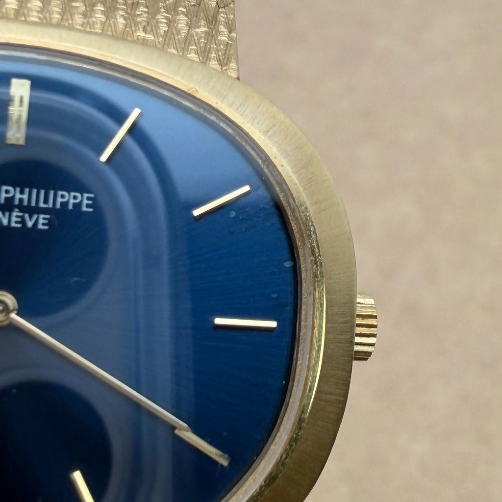 An image of a pre-owned Patek Philippe wristwatch, captured in a close-up view focusing on the right side of the watch face. The angle highlights the gold-toned bezel and crown against a deep blue dial with minimalistic gold hour markers. The watch is positioned slightly tilted, showing the textured band at the top edge.