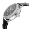 An image of a wristwatch captured from a side profile view, highlighting the crown. The watch features a polished metal case and a black leather strap with visible stitching. The dial is black with white numerals and gold-toned hands. The focus is on the crown, emphasizing its textured design, while the angle provides a clear view of the watch’s thickness and craftsmanship. The image is taken from a close distance, enhancing the detail of the watch's features.
