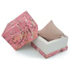 An image of an unworn wristwatch's packaging, featuring a decorative box with a marbled pink and gold pattern. The box is open, revealing a beige cushion inside, meant to hold the wristwatch. The box is positioned at a slight angle, with the lid resting against the side, and the view is taken from a close distance to capture the detailed design.