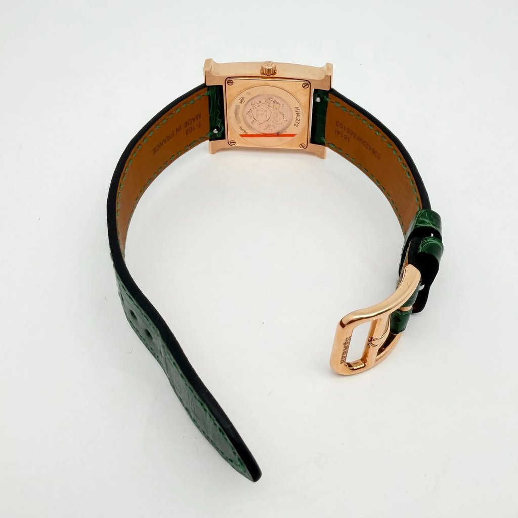 An image of a pre-owned Hermès women's wristwatch displayed from an angled top view. The focus is on the back of the square watch face, showcasing its intricate engravings, with the watch positioned upright on its open green leather strap. The watch is oriented with the buckle slightly off-center to the right, highlighting the gold-toned clasp. The watch is photographed at a close distance against a plain white background.