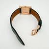 An image of a pre-owned Hermès women's wristwatch displayed from an angled top view. The focus is on the back of the square watch face, showcasing its intricate engravings, with the watch positioned upright on its open green leather strap. The watch is oriented with the buckle slightly off-center to the right, highlighting the gold-toned clasp. The watch is photographed at a close distance against a plain white background.