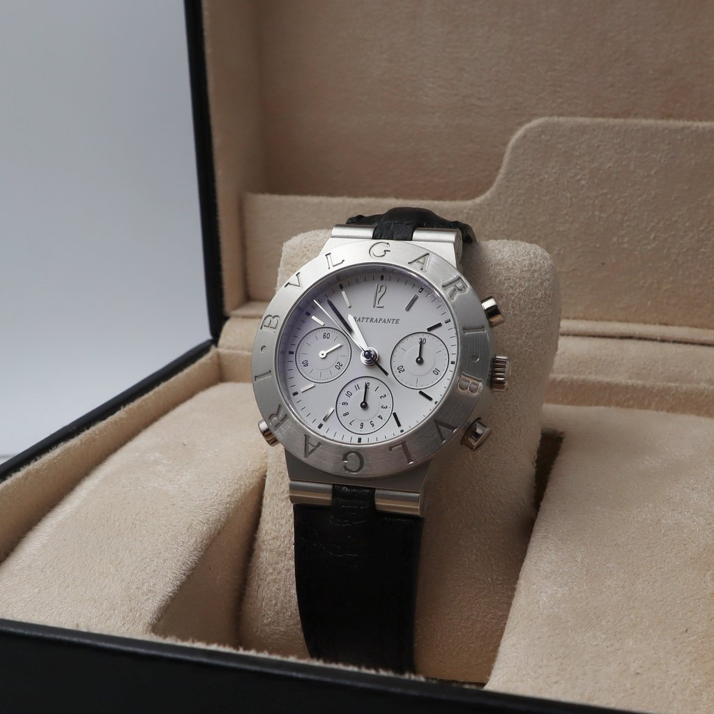 An image of a pre-owned men's Bvlgari wristwatch featuring a chronograph and small seconds subdial. The watch is positioned at a slight angle inside an open beige watch box, showcasing its white dial and black leather strap. The photo is taken from a close distance, emphasizing the engraved Bvlgari logo on the bezel.