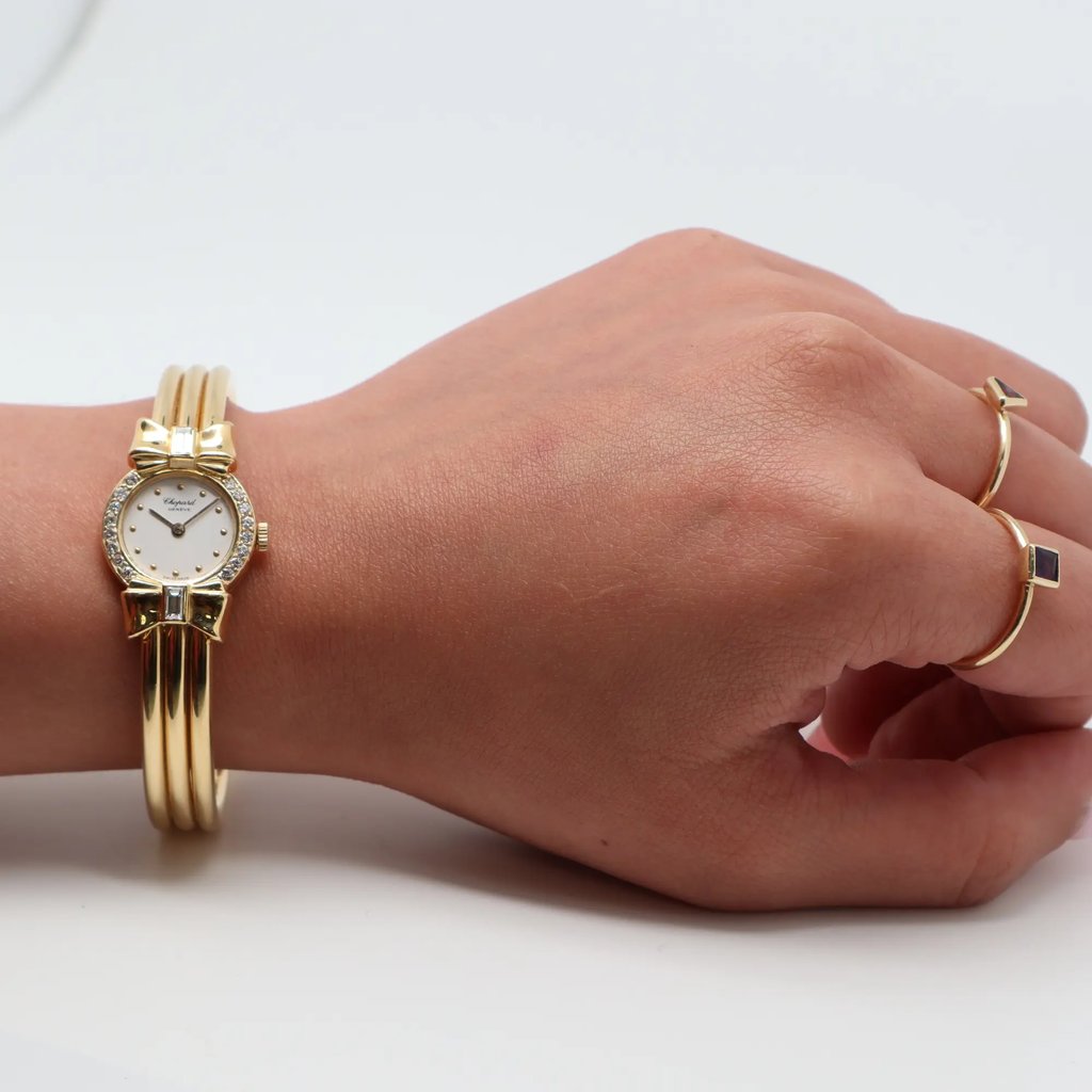 An image of a pre-owned Chopard women's wristwatch on a wrist. The watch features a yellow gold band with a decorative bow design above and below the small, round face. It is positioned horizontally across the wrist, with the face slightly angled towards the camera. The photo is taken from a close distance, highlighting the watch's intricate details and the hand's gold rings.