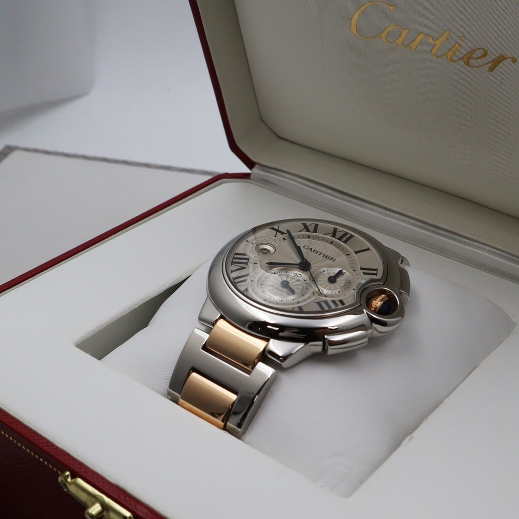 An image of a pre-owned Cartier men's wristwatch with chronograph, date indicator, and small seconds subdial features, positioned at a slight angle on a white cushion inside an open box. The watch has a silver and gold-tone metal bracelet and a round case with a white dial, Roman numerals, and three subdials. The Cartier logo is visible on the top interior part of the box, and the overall image is captured from a top-down perspective with a close-up view, showcasing the watch's details and craftsmanship.