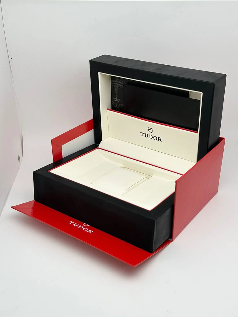An image of a Tudor wristwatch box, shown from a slightly elevated angle. The box is open, revealing a cream interior with the Tudor logo visible. The outer box is black with a red base and flaps. The image is taken from a medium distance, capturing the full view of the box against a plain background.