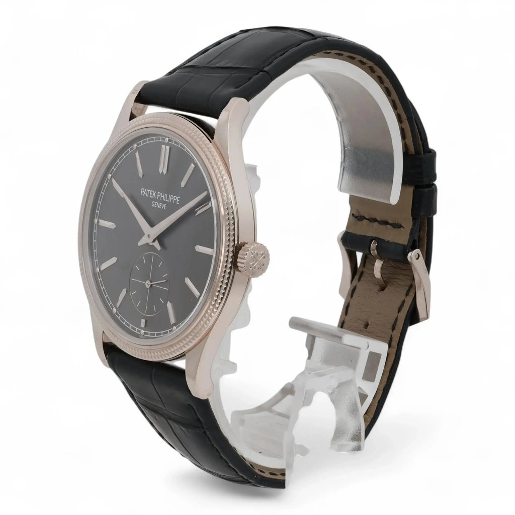 An image of a pre-owned men's Patek Philippe wristwatch, viewed from a slight angle to the left side. The watch has a black leather strap and a dark dial with silver markers. The watch face is positioned prominently in the foreground, with the strap arching upwards in the background. The crown and side details are visible, offering a close-up perspective of the elegant design.