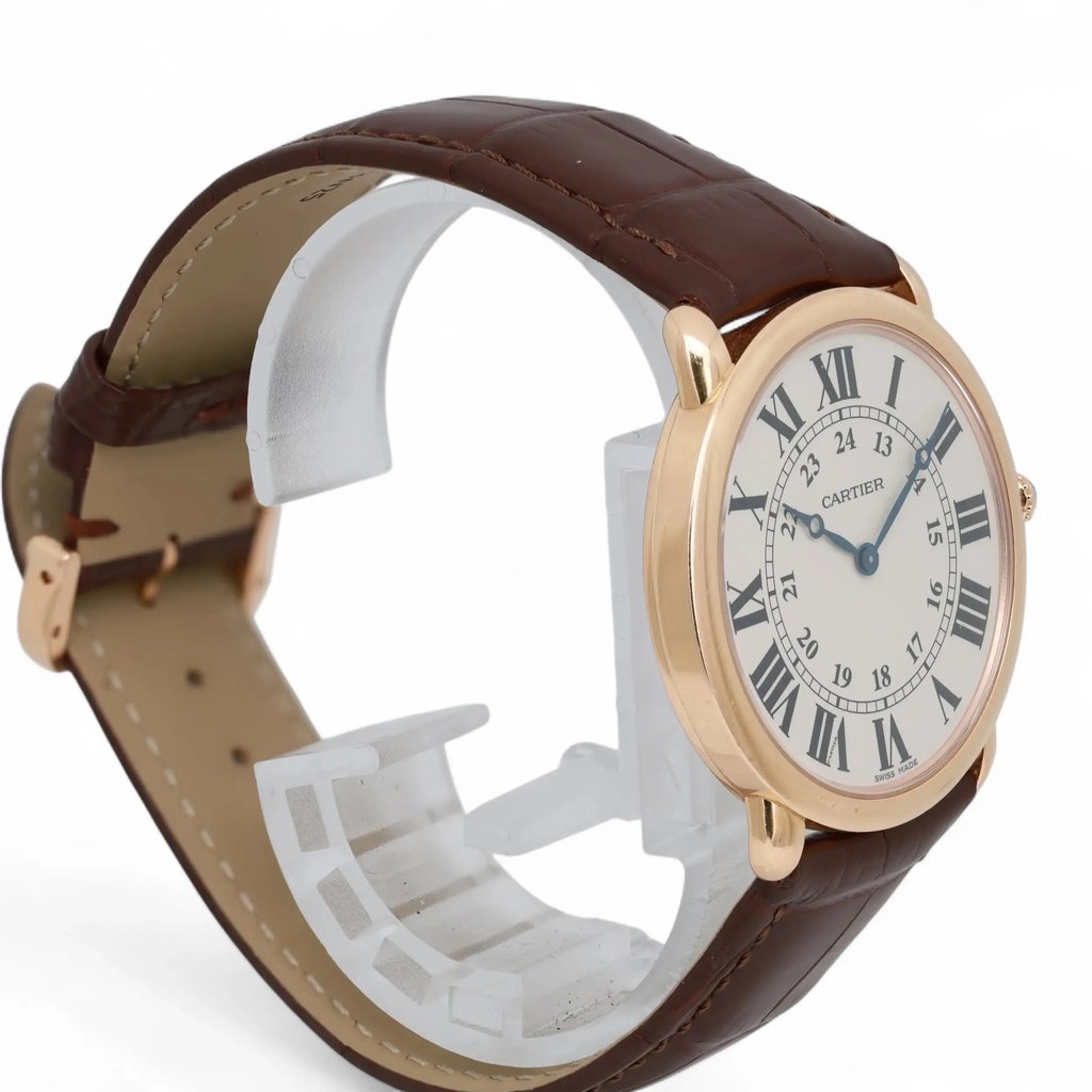 An image of a pre-owned Cartier unisex adult wristwatch. The watch is positioned at a three-quarter angle, slightly elevated, showcasing its face and side. The distance is close, highlighting the details of the watch's design. It features a round, rose gold case and a white dial with black Roman numerals and blue hands. The brown leather strap is visible, with a textured, stitched finish.
