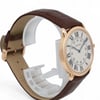 An image of a pre-owned Cartier unisex adult wristwatch. The watch is positioned at a three-quarter angle, slightly elevated, showcasing its face and side. The distance is close, highlighting the details of the watch's design. It features a round, rose gold case and a white dial with black Roman numerals and blue hands. The brown leather strap is visible, with a textured, stitched finish.