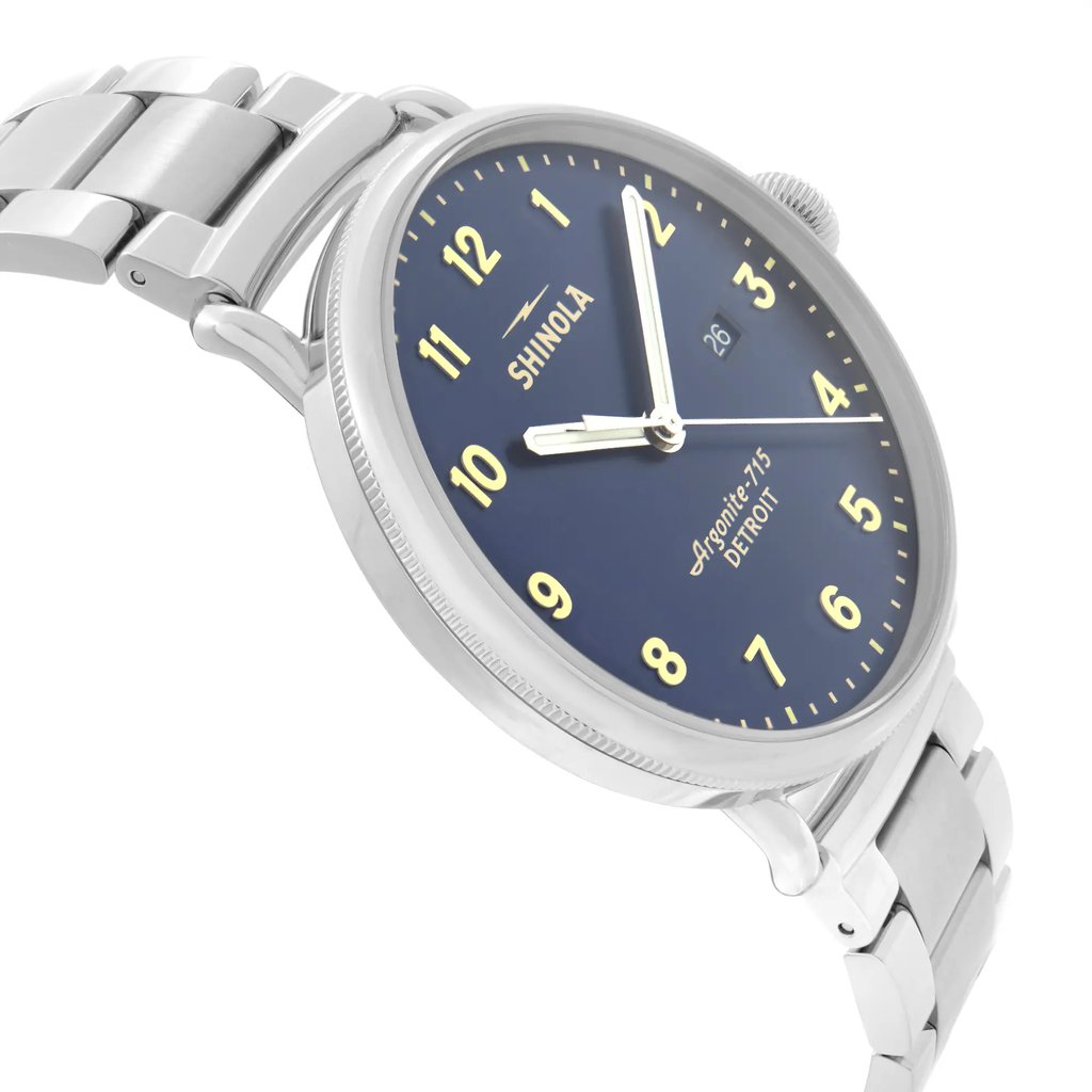 An image of a wristwatch taken from a side profile view opposite the crown, showcasing its sleek design. The watch features a metallic bracelet and a dark blue face with white and yellow markers. The watch is positioned at a slight angle, emphasizing the thickness of the case and the polished links of the bracelet.