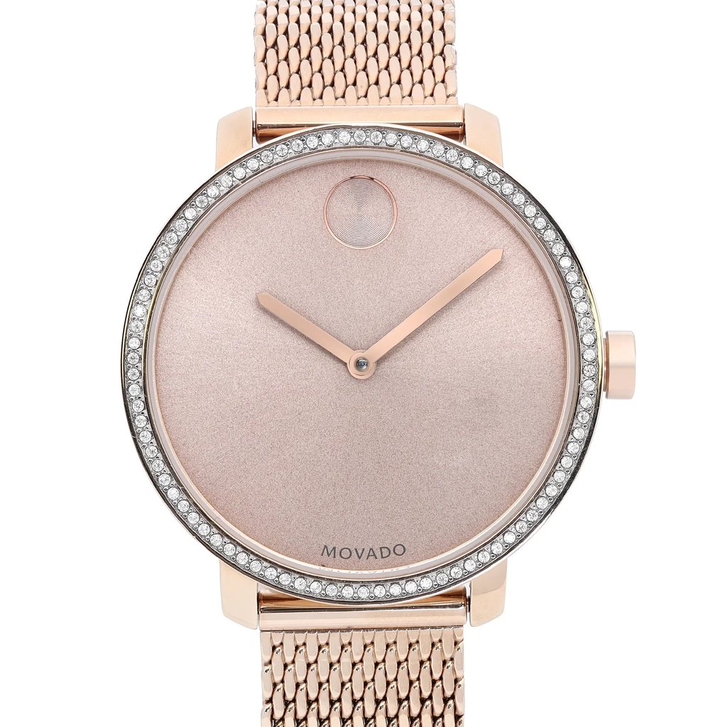 An image of a pre-owned wristwatch taken from a front close-up angle, focusing on the watch face. The watch features a minimalist design with a rose gold-toned dial and matching hands. The bezel is adorned with sparkling stones, and the band has a mesh pattern, also in a rose gold finish. The brand name "Movado" is visible on the dial.