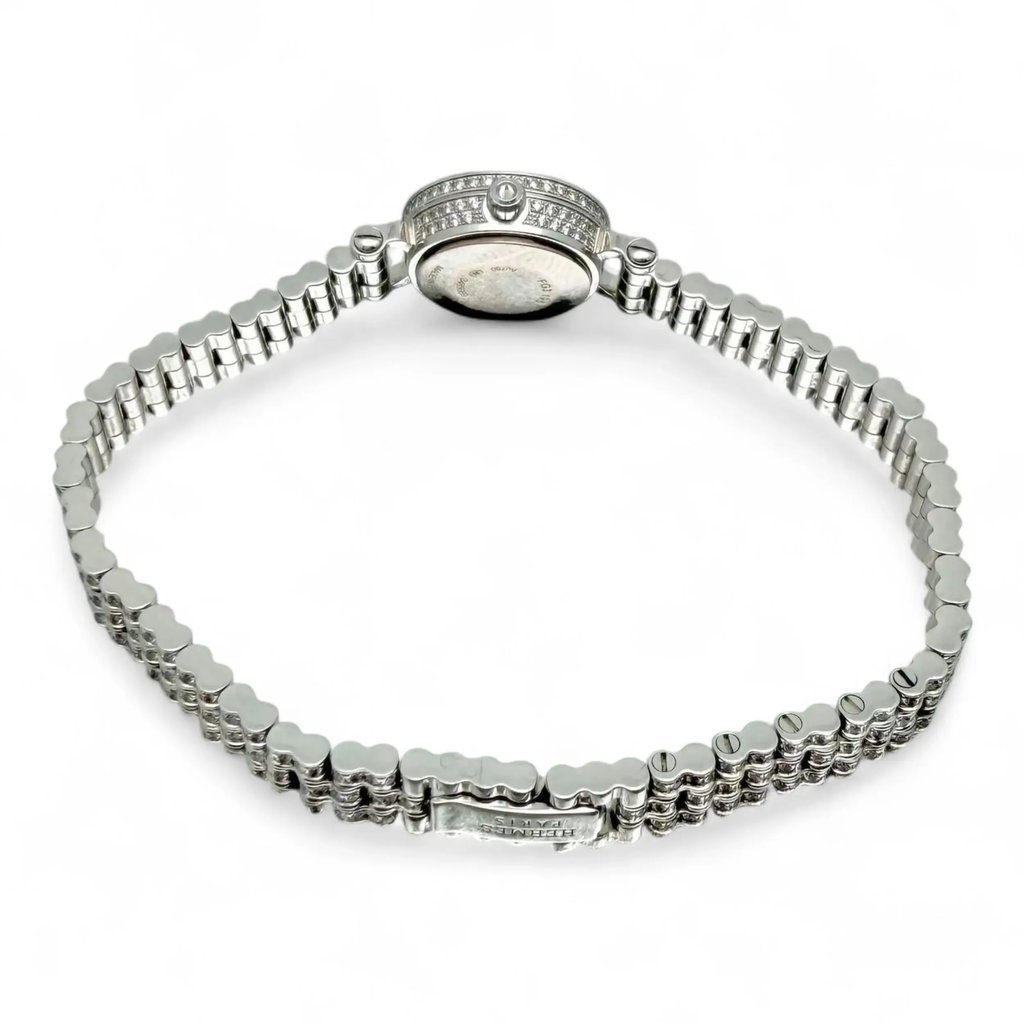 An image of a pre-owned Hermès women's wristwatch displayed from a slightly elevated rear angle. The focus is on the intricate, polished white gold bracelet and the back of the watch case, adorned with small, sparkling details around the edge. The watch is positioned horizontally, with the bracelet forming an arc, showcasing its elegant design.