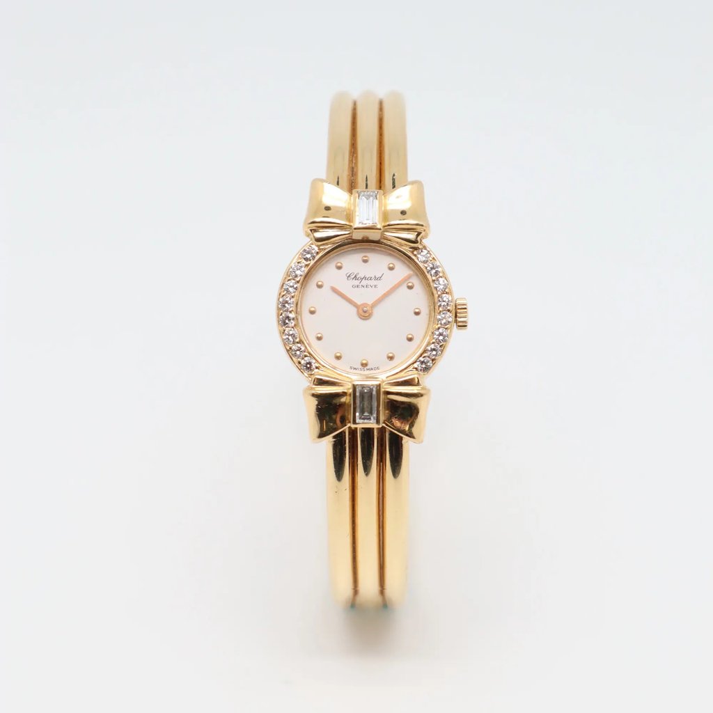 An image of a pre-owned women's Chopard wristwatch, positioned upright with a frontal view. The watch features an 18k yellow gold with a round white face, adorned with a bow design at the top and bottom of the bezel. It is embellished with small diamonds around the dial and has a three-band bracelet. The photo is taken at a close distance, capturing the intricate details of the watch's design.