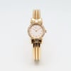An image of a pre-owned women's Chopard wristwatch, positioned upright with a frontal view. The watch features an 18k yellow gold with a round white face, adorned with a bow design at the top and bottom of the bezel. It is embellished with small diamonds around the dial and has a three-band bracelet. The photo is taken at a close distance, capturing the intricate details of the watch's design.