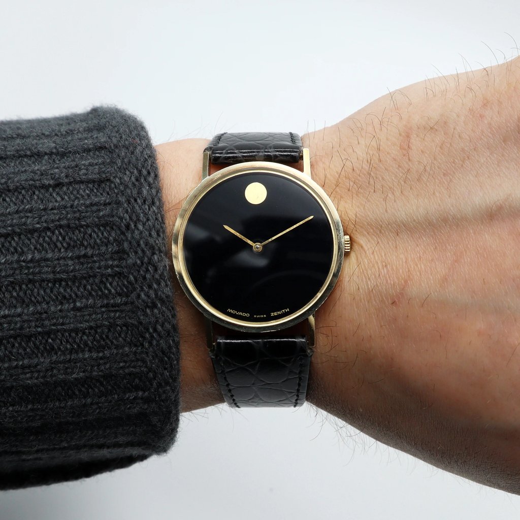 An image of a pre-owned wristwatch viewed from a front full frontal angle. The watch is positioned on a wrist, showing a simple design with a black dial and gold case. The watch features a minimalist face with a single gold dot at the 12 o'clock position and sleek gold hands. It is secured with a black leather strap. The image is taken at a close distance, focusing on the watch's details.