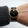 An image of a pre-owned wristwatch viewed from a front full frontal angle. The watch is positioned on a wrist, showing a simple design with a black dial and gold case. The watch features a minimalist face with a single gold dot at the 12 o'clock position and sleek gold hands. It is secured with a black leather strap. The image is taken at a close distance, focusing on the watch's details.