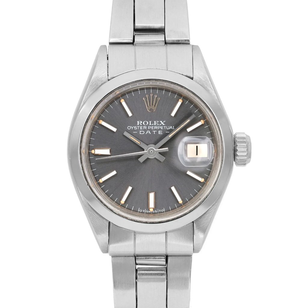 An image of a pre-owned wristwatch, featuring a front close-up shot of the watch face. The watch has a metallic finish with a dark dial, marked with simple hour indices and a date window. The brand's emblem is visible near the 12 o'clock position. The watch is set against a plain background, highlighting its sleek design and craftsmanship.