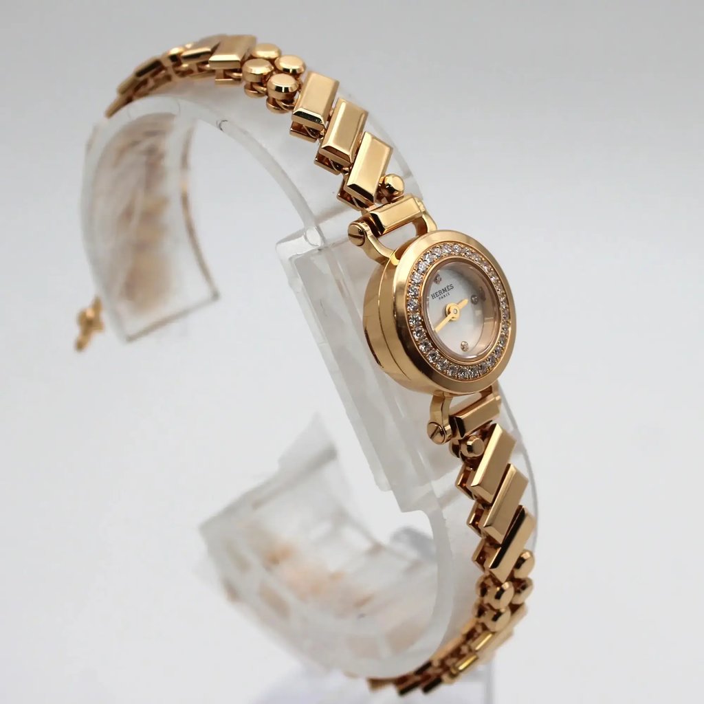 An image of a pre-owned women's HERMÈS wristwatch, positioned at a slight angle to the right. The watch is displayed on a transparent stand, highlighting its rose gold bracelet and diamond-encrusted bezel. The photo is taken from a close-up perspective, emphasizing the luxurious details of the timepiece.