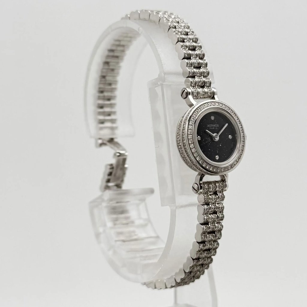 An image of a pre-owned Hermès women's wristwatch positioned at a slight angle, showcasing the elegant design. The watch features a round face with a dark dial, set within a bezel adorned with small diamonds. The white gold, textured bracelet is prominently displayed as it curves around a transparent stand, highlighting the craftsmanship from a close-up perspective.