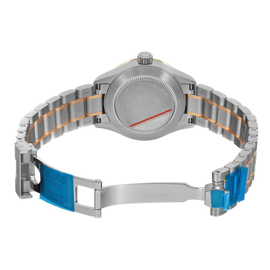 An image of an unworn wristwatch showing the back view with an open clasp. The watch is positioned upright, displaying the case back and the metal bracelet. The bracelet features a combination of silver and gold tones, with blue protective stickers on the clasp and links. The angle is slightly elevated, providing a clear view of the back of the watch and the open clasp from a close distance.