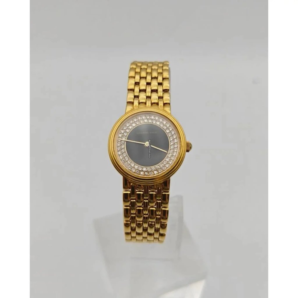 An image of a pre-owned women's Audemars Piguet wristwatch displayed vertically. The watch features a yellow gold band with a woven link design and a circular face. The dial is onyx with a surrounding ring of sparkling diamonds. The watch is positioned at a slight angle, resting on a clear stand, and photographed from a medium distance against a neutral background.